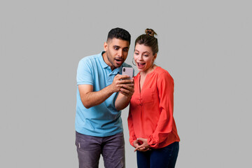 Friends reading shocking news! Portrait of amazed attractive young couple in casual wear reading unexpected message with open mouth, using mobile phone. isolated on gray background indoor studio shot