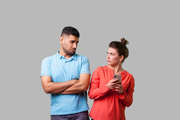 Portrait of funny resentful man in casual wear standing with crossed arms, looking disapprovingly at woman holding cellphone, addicted to internet. isolated on gray background, indoor studio shot