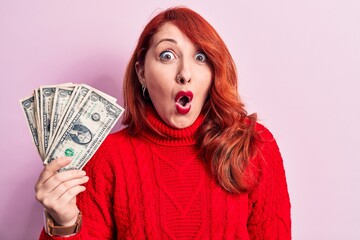 Young beautiful redhead woman holding bunch of dollars banknotes over pink background scared and amazed with open mouth for surprise, disbelief face