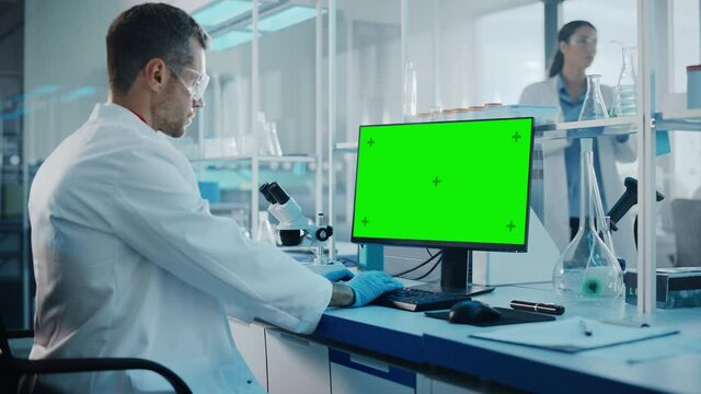 Medical Science Laboratory: Microbiologist Working on Computer with Display with Green Chroma Key Screen. Diverse Multi-Ethnic Team of Biotechnology Scientists Developing Drugs. Moving Shot