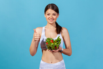 Healthy positive woman in white sportswear with perfect slim body showing thumbs up holding big bowl with fresh vegetable salad, keeping detox diet. Indoor studio shot isolated on blue background