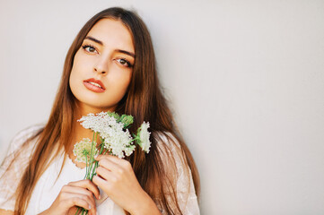 Fashion portrait of beautiful young woman posing next to white wall, holding bluthenball flowers