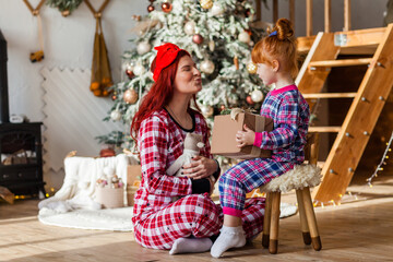 Happy woman and kid wearing checkered pajamas is sitting beside the Christmas tree open presents, winter holidays for mother and daughter