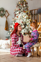 Happy woman and kid wearing checkered pajamas is sitting beside the Christmas tree open presents, winter holidays for mother and daughter