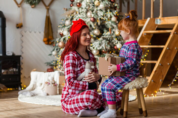Obraz na płótnie Canvas Happy woman and kid wearing checkered pajamas is sitting beside the Christmas tree open presents, winter holidays for mother and daughter