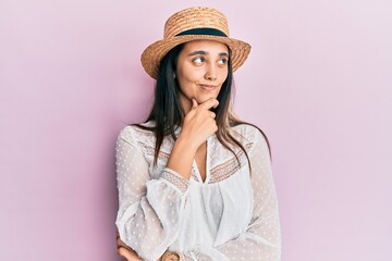 Young hispanic woman wearing summer hat thinking concentrated about doubt with finger on chin and...
