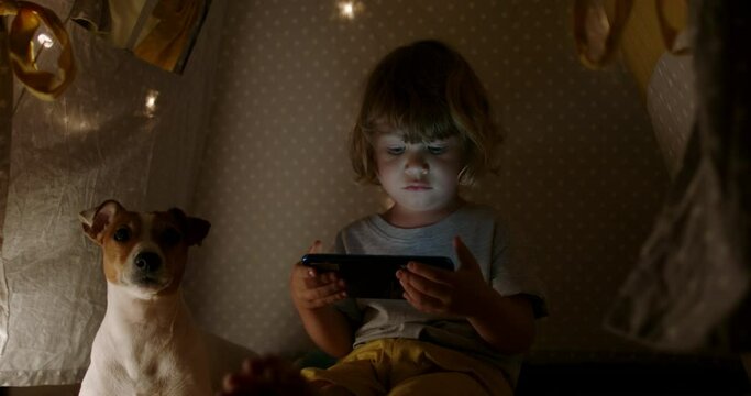 Cute funny caucasian baby boy is watching cartoons on a mobile phone while sitting in cozy tent with string lights 4k footage
