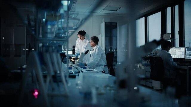 Modern Medical Research Laboratory: Two Scientists Working, Using Digital Tablet, Analyzing Test, Talking. Advanced Scientific Pharmaceutical Lab for Medicine, Biotechnology Development. Evening Time