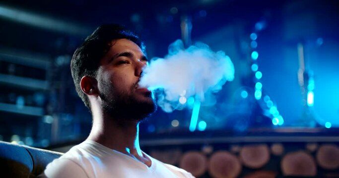 Closeup shot of bearded asian guy smoking a shisha in nightclub, exhaling clouds of smoke in neon light, relaxing - nightlife, niccotine addiaction concept 4k footage portrait