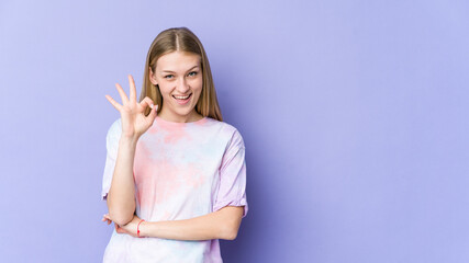 Young blonde woman isolated on purple background winks an eye and holds an okay gesture with hand.