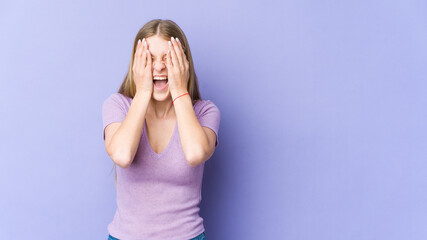 Young blonde woman isolated on purple background having fun covering half of face with palm.