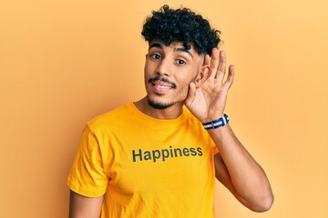 Fototapeta na wymiar Young arab handsome man wearing tshirt with happiness word message smiling with hand over ear listening and hearing to rumor or gossip. deafness concept.