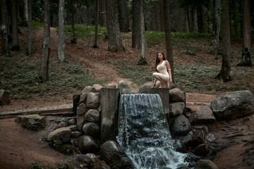A young woman in a macrame dress is sitting on a large fountain in a green forest. The concept of balance and natural cosmetics.