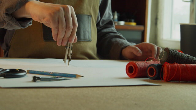 A tailor in an apron makes a marking with compasses on the pattern. In the foreground, reels with colored threads. - Powered by Adobe