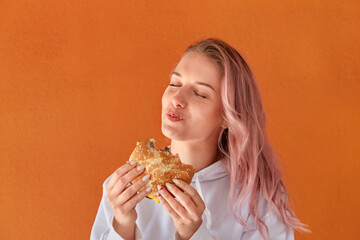 A young woman with pink hair eats fast food with relish. Orange background. The concept of junk food.