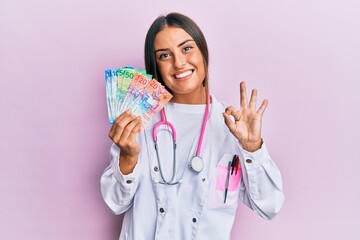 Beautiful hispanic woman wearing medical uniform holding swiss franc banknotes doing ok sign with fingers, smiling friendly gesturing excellent symbol