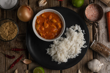 Top view of plate with delicious chicken tikka masala curry and rice on rustic wooden table with spices background. Traditional Indian dish. Exotic tasty meal. Appetizing photo for menu or cookbook.