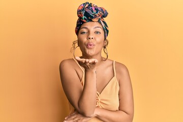 Young hispanic girl wearing hair turban over yellow background looking at the camera blowing a kiss with hand on air being lovely and sexy. love expression.