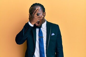 Handsome young black man wearing business suit and tie peeking in shock covering face and eyes with hand, looking through fingers afraid