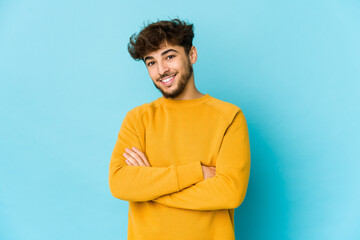 Young arab man on blue background who feels confident, crossing arms with determination.