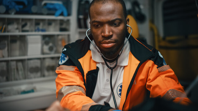 Close-up Portrait Shot Of A Serious And Focused Black African American Paramedic In An Ambulance Vehicle With An Injured Patient. Emergency Medical Technician Uses Stethoscope To Monitor The Condition