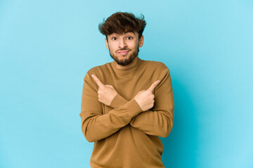 Young arab man on blue background points sideways, is trying to choose between two options.