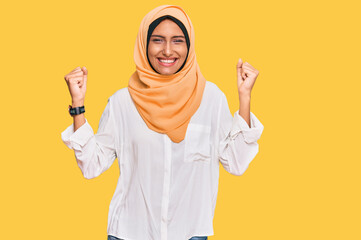 Young brunette woman wearing traditional islamic hijab scarf screaming proud, celebrating victory and success very excited with raised arms