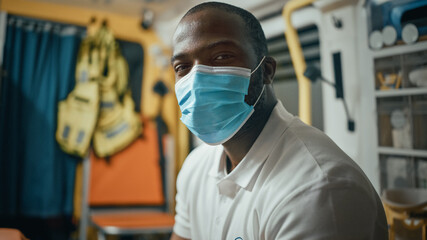 Fototapeta na wymiar Calm Black African American EMS Professional Paramedic Looks at Camera While Wearing a Safety Face Mask in Ambulance Vehicle. Successful Emergency Medical Technician Outside the Healthcare Hospital.
