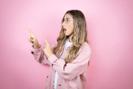Young beautiful blonde woman with long hair standing over pink background surprised and pointing her fingers side