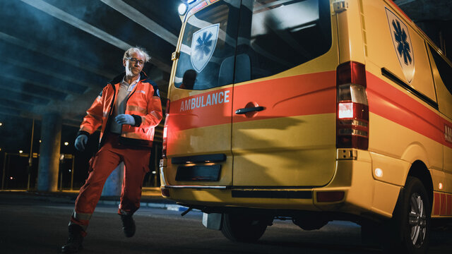 Team Of EMS Paramedics Quickly Take Out A Stretcher From Ambulance Vehicle. Emergency Care Assistants Arrived On The Scene Of A Traffic Accident On A Street At Night And Run To Open Back Doors.