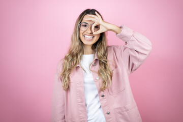 Fototapeta premium Young beautiful blonde woman with long hair standing over pink background doing ok gesture shocked with smiling face, eye looking through fingers