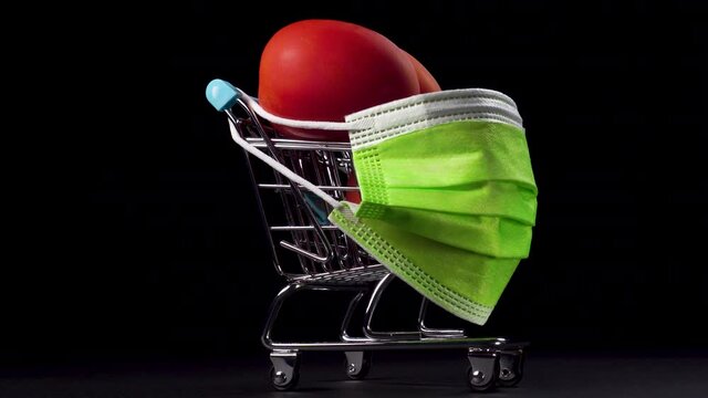 Supermarket cart with red tomatoes wears a bright green medical protective mask against a black background. Close-up. Miniature.
