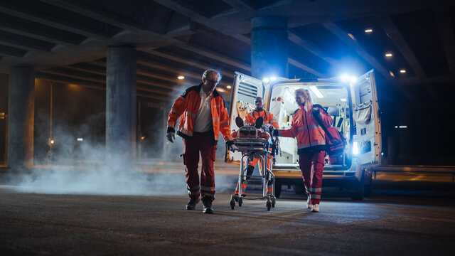 Team Of EMS Paramedics React Quick To Bring Injured Patient To Healthcare Hospital And Get Him Out Of Ambulance On A Stretcher. Emergency Care Assistants Help Young Man To Stay Alive After Accident.