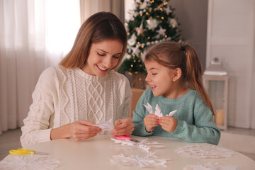 Fototapeta premium Happy mother and daughter making paper snowflakes at table near Christmas tree indoors