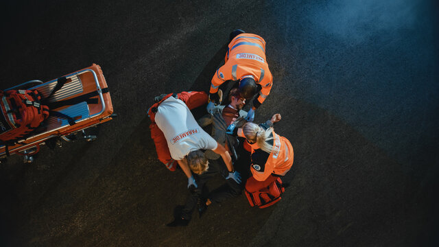 Aerial Portrait Of A Young Injured Man Involved In Traffic Accident Is Being Saved By Medical Team Of EMS Paramedics On The Street At Night. Emergency Care Assistants Provide Essential First Aid Help.