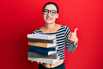Young caucasian woman wearing glasses and holding books smiling happy and positive, thumb up doing excellent and approval sign