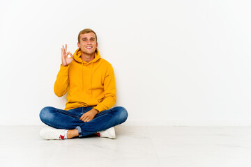 Fototapeta na wymiar Young caucasian man sitting on the floor cheerful and confident showing ok gesture.