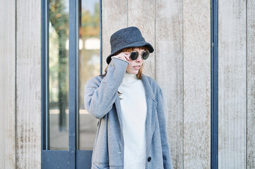 Portrait young pretty Caucasian girl with sunglasses, dressed in a gray jacket and a white sweater and a hat.