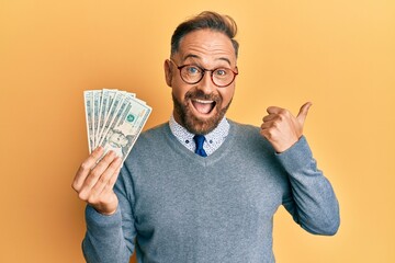 Handsome middle age man holding 20 dollars banknotes pointing thumb up to the side smiling happy with open mouth