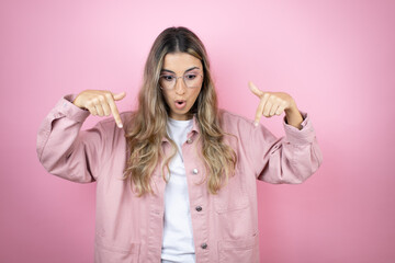Young beautiful blonde woman with long hair standing over pink background surprised, looking down and pointing down with fingers and raised arms