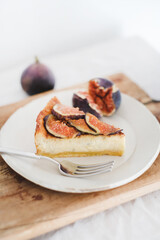 Slice of homemade gluten free fig cheesecake with fresh seasonal figs pieces on white plate over rustic wooden board, white wall at background. Healthy dessert, artisan sweet food concept