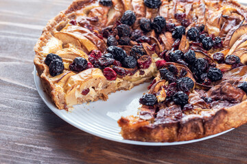 Open shortcrust apple pie with cranberry, raisins, cinnamon without one piece on white plate on rustic wooden table.  Close up side view.