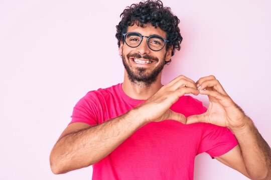 Handsome young man with curly hair and bear wearing casual clothes and glasses smiling in love showing heart symbol and shape with hands. romantic concept.