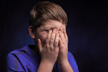 caucasian teenage boy with light brown hair, wearing a purple sports t-shirt, covering his face with his hands on an isolated background