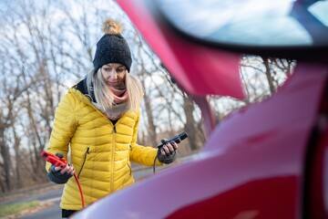 Beautiful blonde woman in yellow jacket trying to start her broken car with jumper cables.