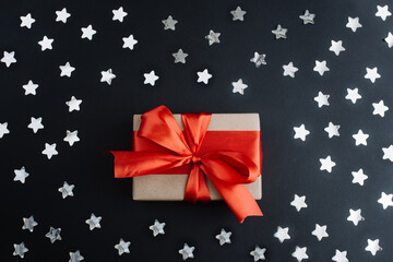  Present box with red bow on black background with sparkling confetti. Flat lay, top view, copy space.