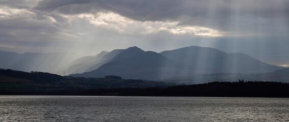 Sunlight filtering through clouds over the Menai Straits with Snowdonia mountain range behind. Wintry landscape in monochrome colours.