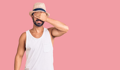 Young hispanic man wearing casual summer hat smiling and laughing with hand on face covering eyes for surprise. blind concept.