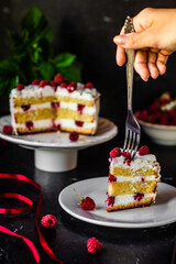 A fork in the piece of tasty biscuit sponge cake with cream cheese and fresh raspberries and mint leaves. Sweet dessert with berries on a dark table, low key, negative space, vertical orientation