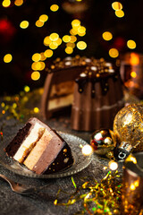 A piece of tasty Three Chocolates mousse cake with ganache in Christmas and New Year decorations. Chocolate cake with layers. Bokeh lights background. Vertical orientation. Christmas Eve. Low Key.